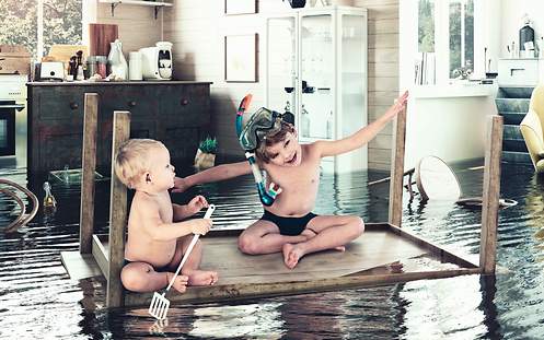 Zwei Kinder schwimmen auf einem Tisch durch die Küche, vorbei an Hausgeräten. 