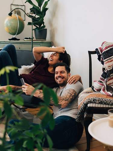 Ein Pärchen sitzt lachend im Wohnzimmer, weil es mit einer Kombi aus Hausrat, Haftpflicht und Rechtsschutz versichert ist.