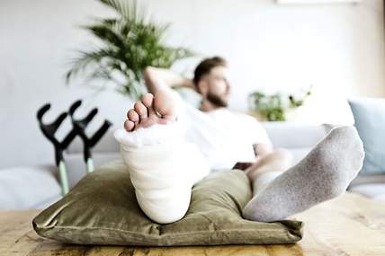 Ein Mann liegt auf einer Couch. Sein rechtes Bein ist eingegipst, an der Wand lehnt ein Paar Krücken.