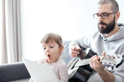 Das Bild zeigt einen Mann mit Gitarre mit seiner Tochter vor dem Laptop