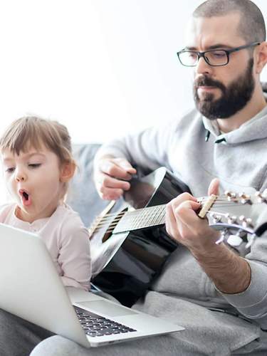 Das Bild zeigt einen Mann mit Gitarre mit seiner Tochter vor dem Laptop