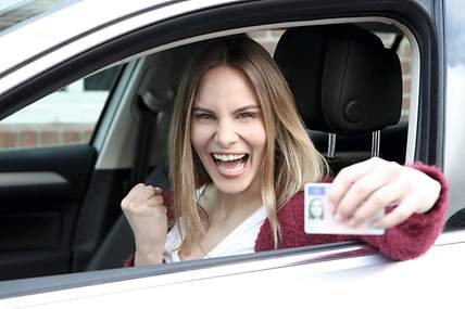 Eine Fahrschülerin und ihr Fahrlehrer sitzen nach bestandener Führerscheinprüfung im Auto und zeigen einen Daumen nach oben.