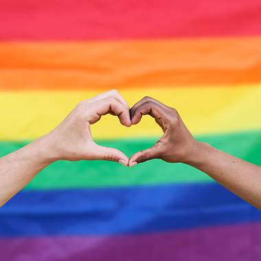 Zwei Hände bilden ein Herz vor einer Regenbogenfarbenflagge
