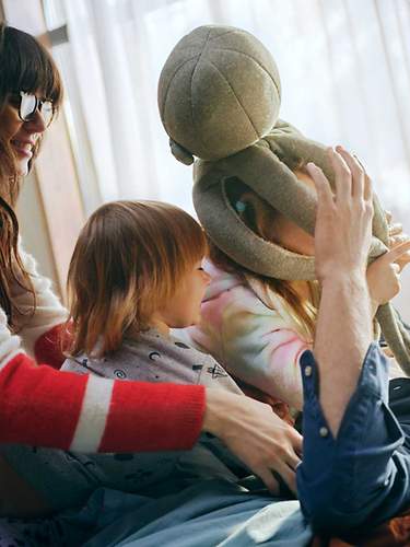 Das Bild zeigt eine junge Familie, die auf einem Bett mit einem Stofftier herumalbert.