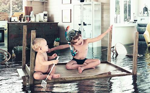 Elektronische Geräte in der Hausratversicherung Zwei Kinder schwimmen auf einem Tisch durch die Küche, vorbei an Hausgeräten.