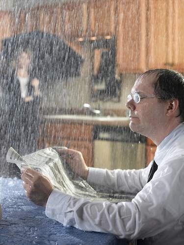 Ein Mann sitzt an einem Esstisch, während es reinregnet. Er fragt sich, welche Versicherung für den Schaden aufkommt.