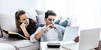 Ein junges Paar sitzt mit Versicherungsunterlagen, einem Laptop und Taschenrechner im Wohnzimmer.