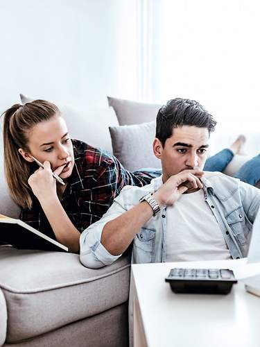 Ein junges Paar sitzt mit Versicherungsunterlagen, einem Laptop und Taschenrechner im Wohnzimmer.