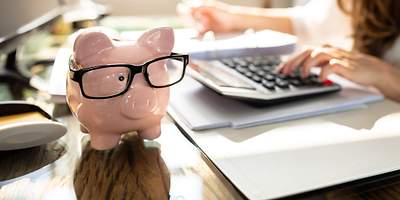 Sparschwein mit Brille steht auf einem Tisch, eine Frau berechnet, wie viel man mit einer günstigen Versicherung sparen kann.