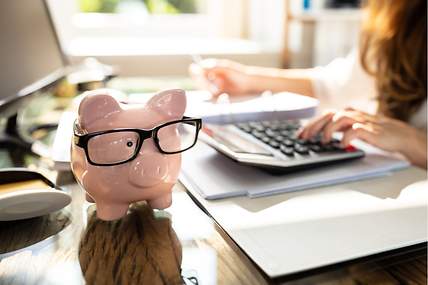 Sparschwein mit Brille steht auf einem Tisch, eine Frau berechnet, wie viel man mit einer günstigen Versicherung sparen kann.