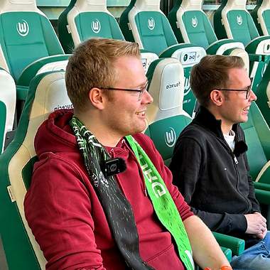 Zwei Männder sitzen auf einer Tribüne im Fußballstadion und freuen sich. 