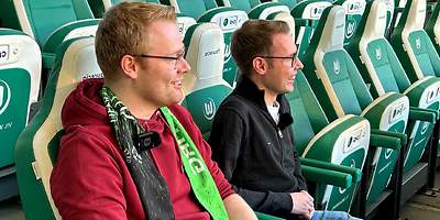 Zwei Männder sitzen auf einer Tribüne im Fußballstadion und freuen sich. 