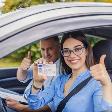 Eine Fahranfängerin hält Ihren Führerschein hoch und freut sich. Ihr Fahrlehrer sitzt daneben.