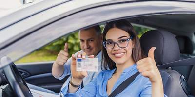 Eine Fahranfängerin hält Ihren Führerschein hoch und freut sich. Ihr Fahrlehrer sitzt daneben.