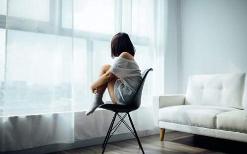 Eine Frau sitzt mit angewinkelten Knien auf einem Stuhl und sieht aus dem Fenster